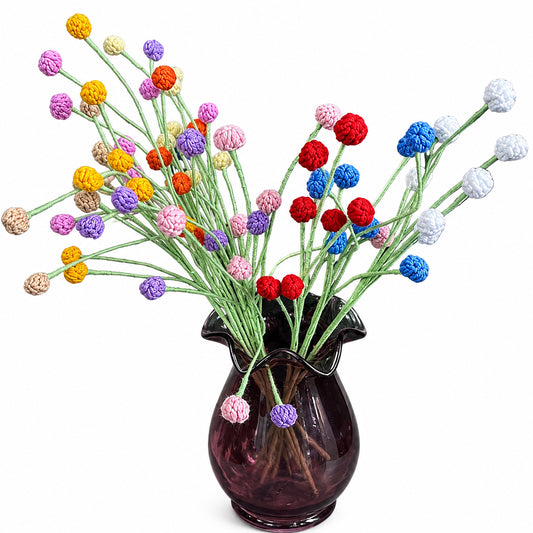 A glass vase filled with artificial flower stems, featuring a variety of colorful pom-pom blossoms in red, blue, purple, pink, yellow, orange, and white on green stems, set against a plain white background.