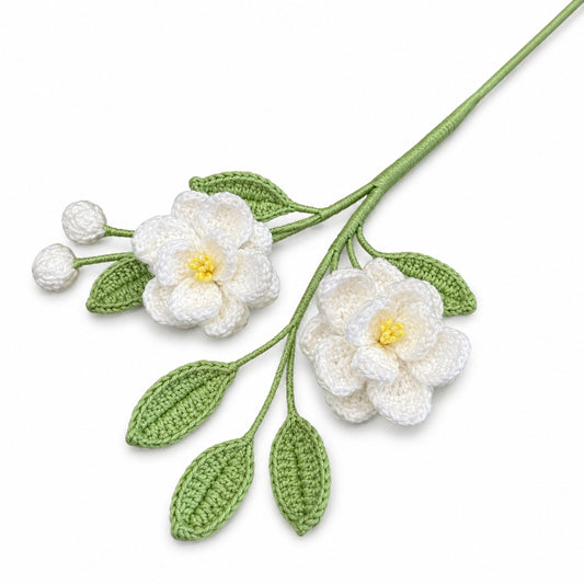 Crocheted floral branch with two white flowers, yellow centers, green leaves, and white buds on a green stem, photographed on a clean white background.