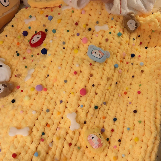 Yellow knitted blanket with cartoon characters and colorful dots, person holding a phone in the background.