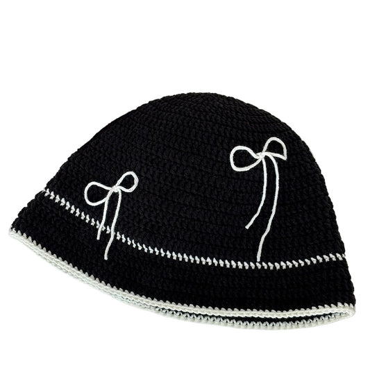 Black crochet hat with white bow patterns on a white surface, next to yarn and a newspaper.