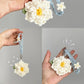 Hand holding a white crocheted flower keychain with a blue strap against a gray background