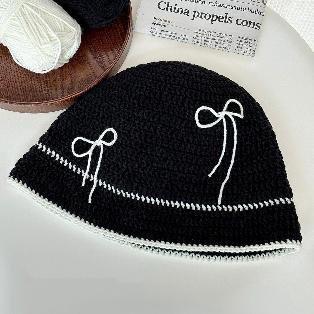 Black crochet hat with white bow patterns on a white surface, next to yarn and a newspaper.