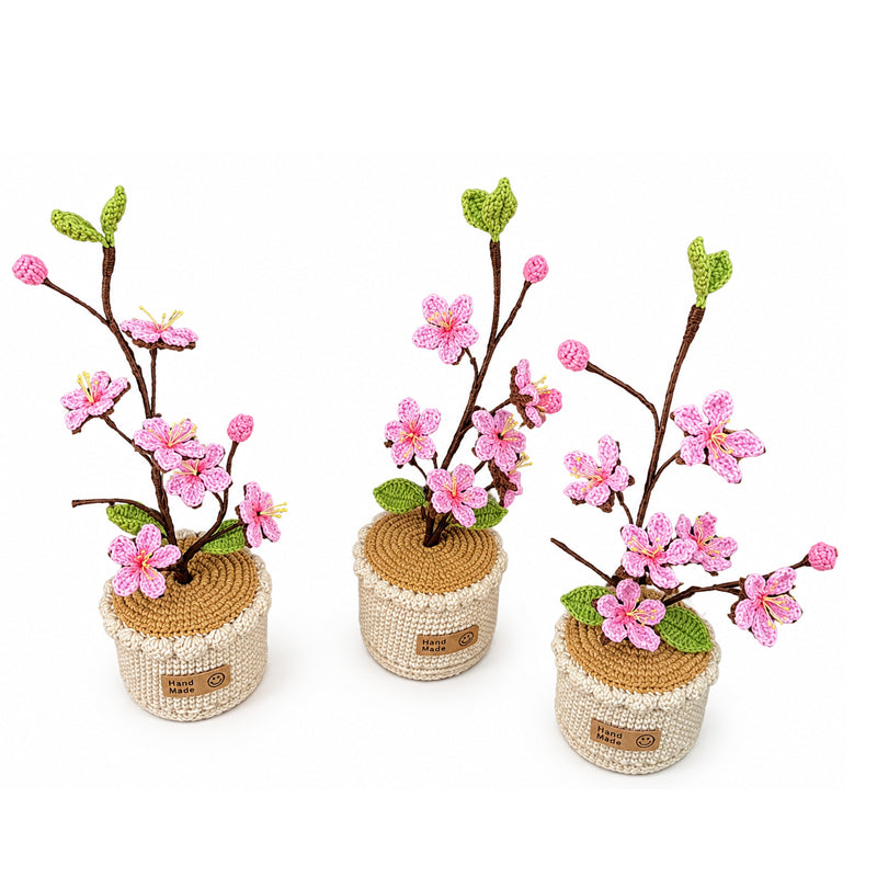 Three handmade crocheted cherry blossom planties with pink flowers, green leaves, and brown branches, arranged on a white background.