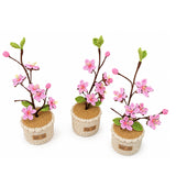 Three handmade crocheted cherry blossom planties with pink flowers, green leaves, and brown branches, arranged on a white background.