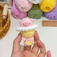 Hand holding a small crocheted cupcake with pink frosting and a bow, with yarn balls in the background.