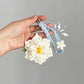 Hand holding a white crocheted flower keychain with a blue string against a gray background