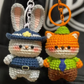 Two crochet keychains, one resembling a rabbit in a blue outfit and the other resembling a cat in an orange outfit, on a dark background.