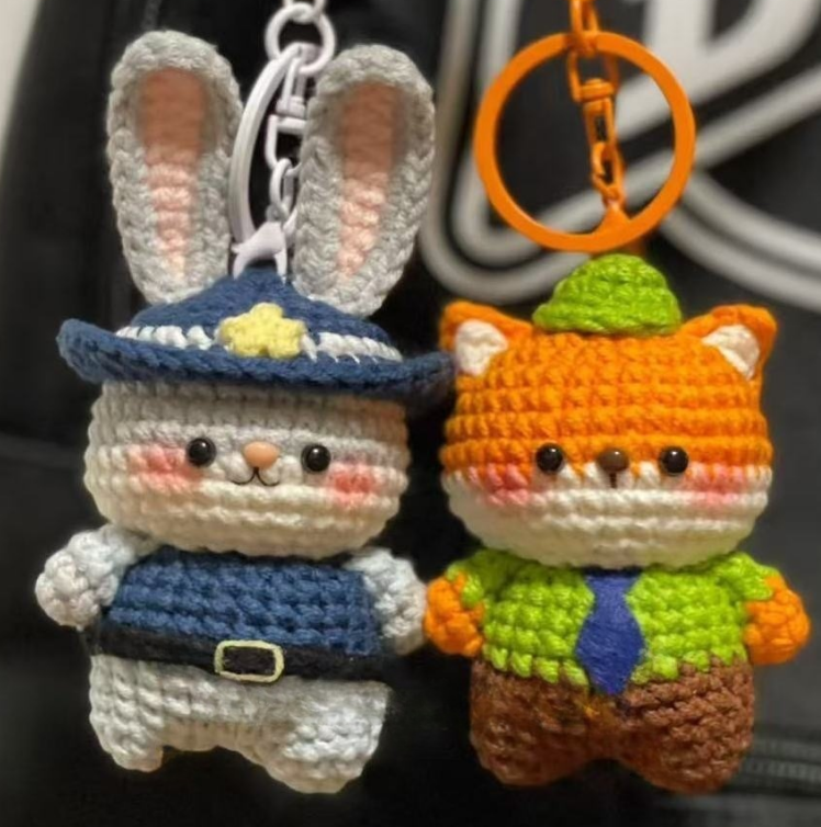 Two crochet keychains, one resembling a rabbit in a blue outfit and the other resembling a cat in an orange outfit, on a dark background.