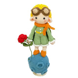 Crocheted character with a red rose, wearing sunglasses and an orange scarf, on a white surface.
