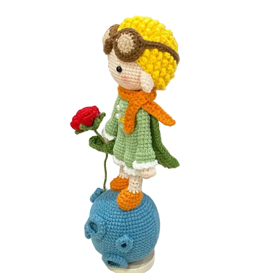 Crocheted character with yellow hair, goggles, and a green outfit holding a red rose on a white surface.