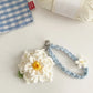 Crocheted flower keychain with a blue strap on a light surface next to yarn and a red label.