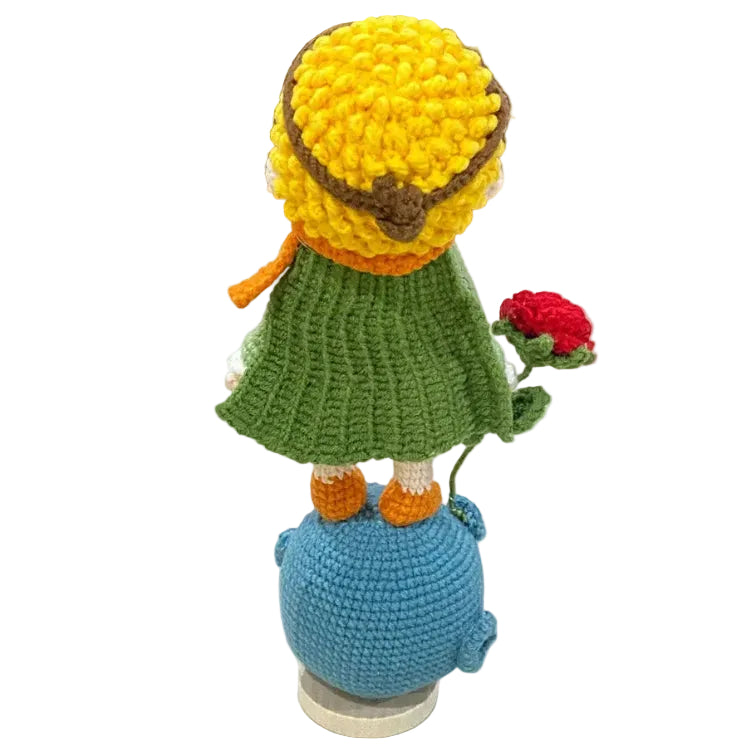 Crocheted character with a yellow hat, green dress, and blue pot on a white surface.