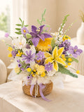 A woven basket floral arrangement featuring purple and yellow crocheted flowers, green foliage, white tulle wrapping, and a lavender ribbon, displayed on a light beige table in a softly lit indoor setting.