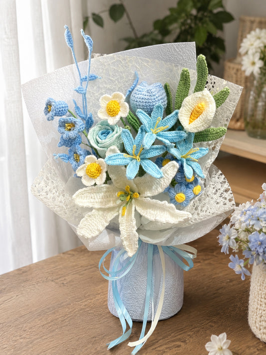 Handmade crocheted flower bouquet in light blue, white, and green, wrapped in white lace paper and tied with a light blue ribbon, displayed on a wooden table.