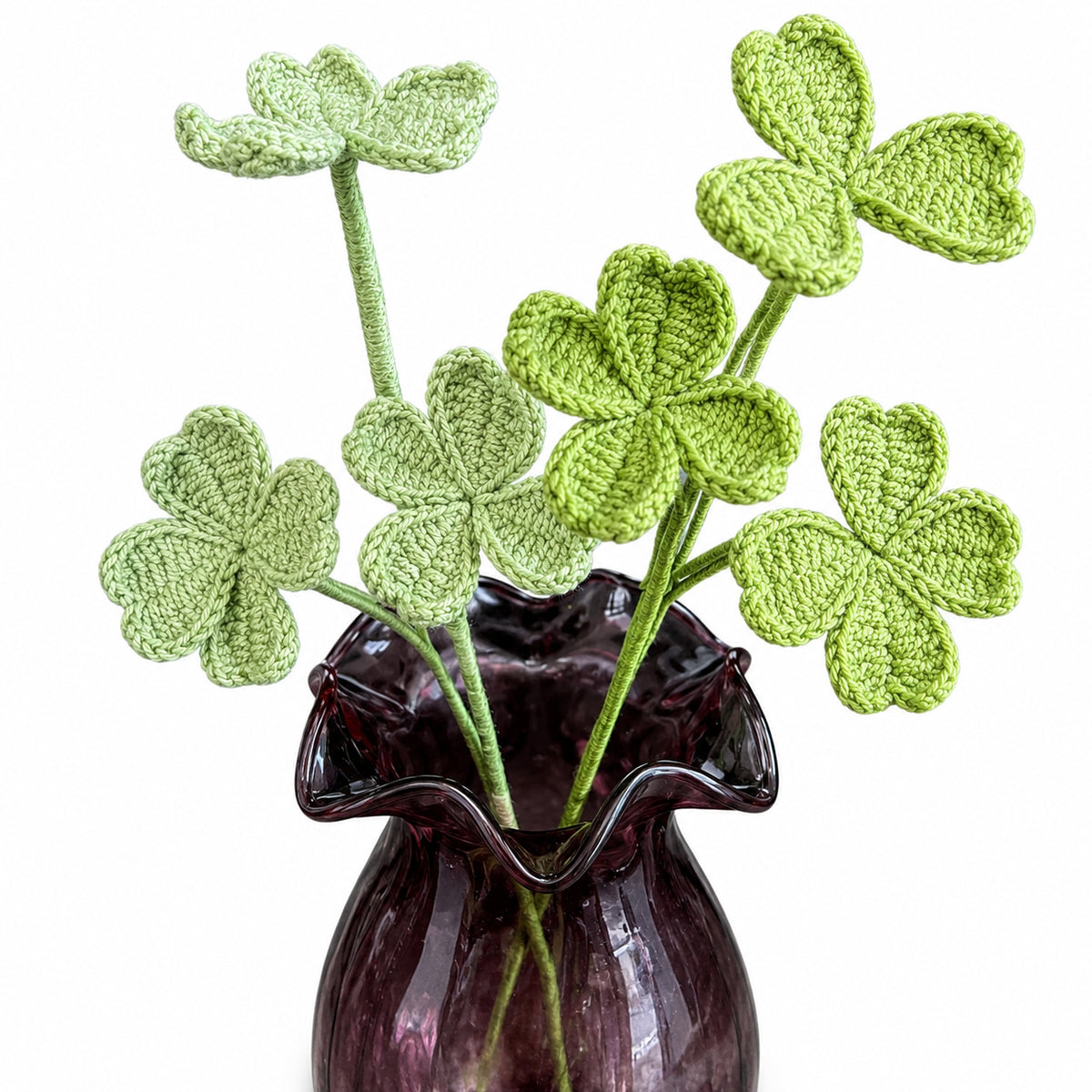 Five green crocheted shamrock flowers with long stems arranged in a dark purple ruffled glass vase against a plain white background.