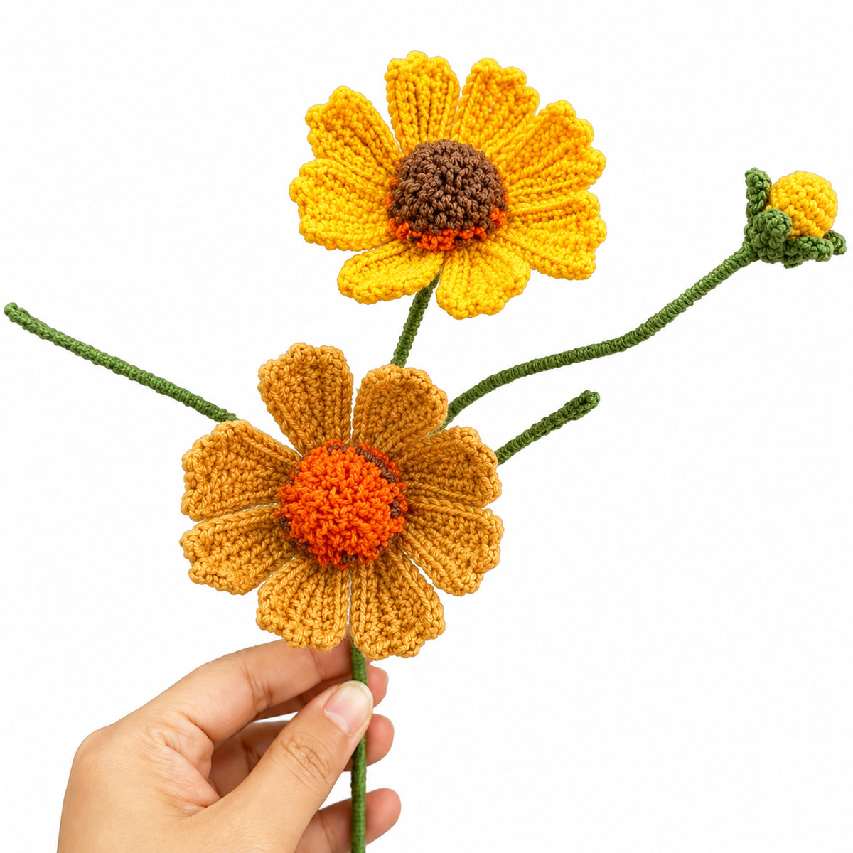 Two crocheted daisy-like flowers with green stems and a small matching bud, held against a white background. One flower is held by a human hand, so this is classified as Lifestyle under the schema rules.