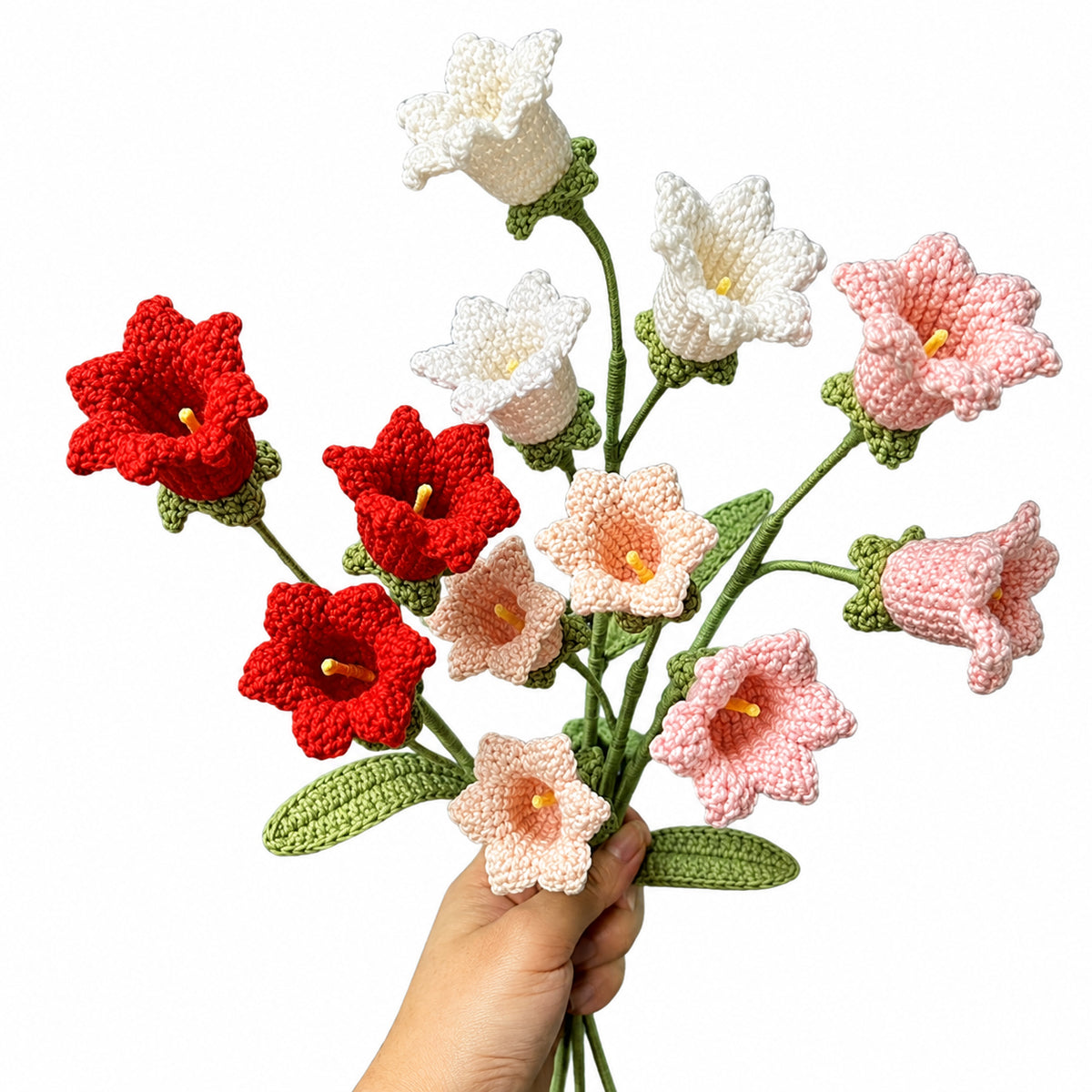 A hand holding a bouquet of crocheted bell-shaped flowers in red, white, and light pink, with green stems and leaves, against a plain white background.