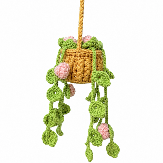 A crocheted hanging planter with a mustard yellow basket, green leafy vines, and pink flowers, suspended against a plain white background.