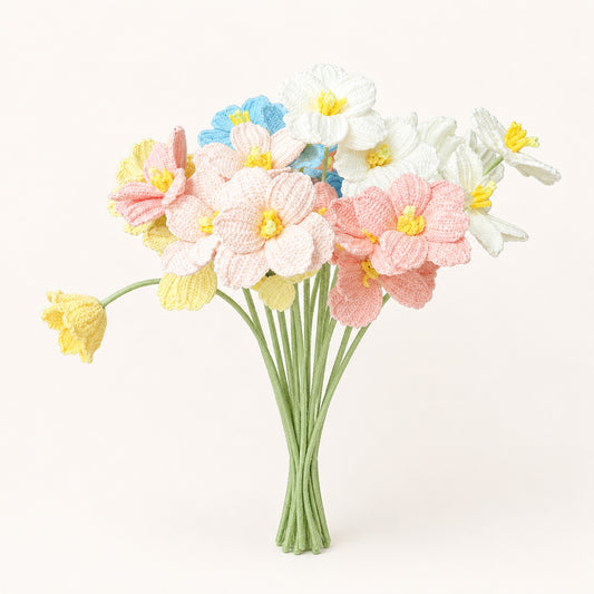 A bouquet of crocheted spring flowers, including pink daffodils, white daisies, yellow buttercups, and a blue flower, arranged in a bundle of green stems against a plain off-white background.