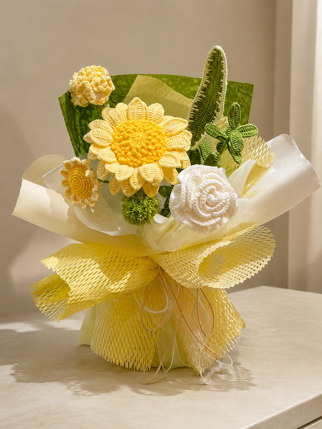 A handmade crochet flower bouquet featuring yellow daisies, a white rose, green cactus-like stems, and textured yellow wrapping paper, displayed on a light beige surface.