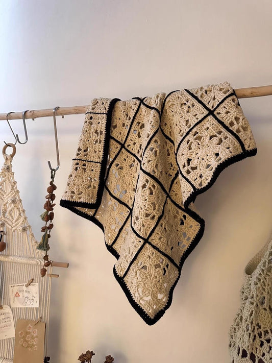 Crocheted blanket with black trim hanging on a wooden rack against a neutral wall.