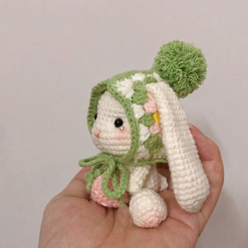 Hand holding a small crochet toy with a green hat and pink accents against a plain background