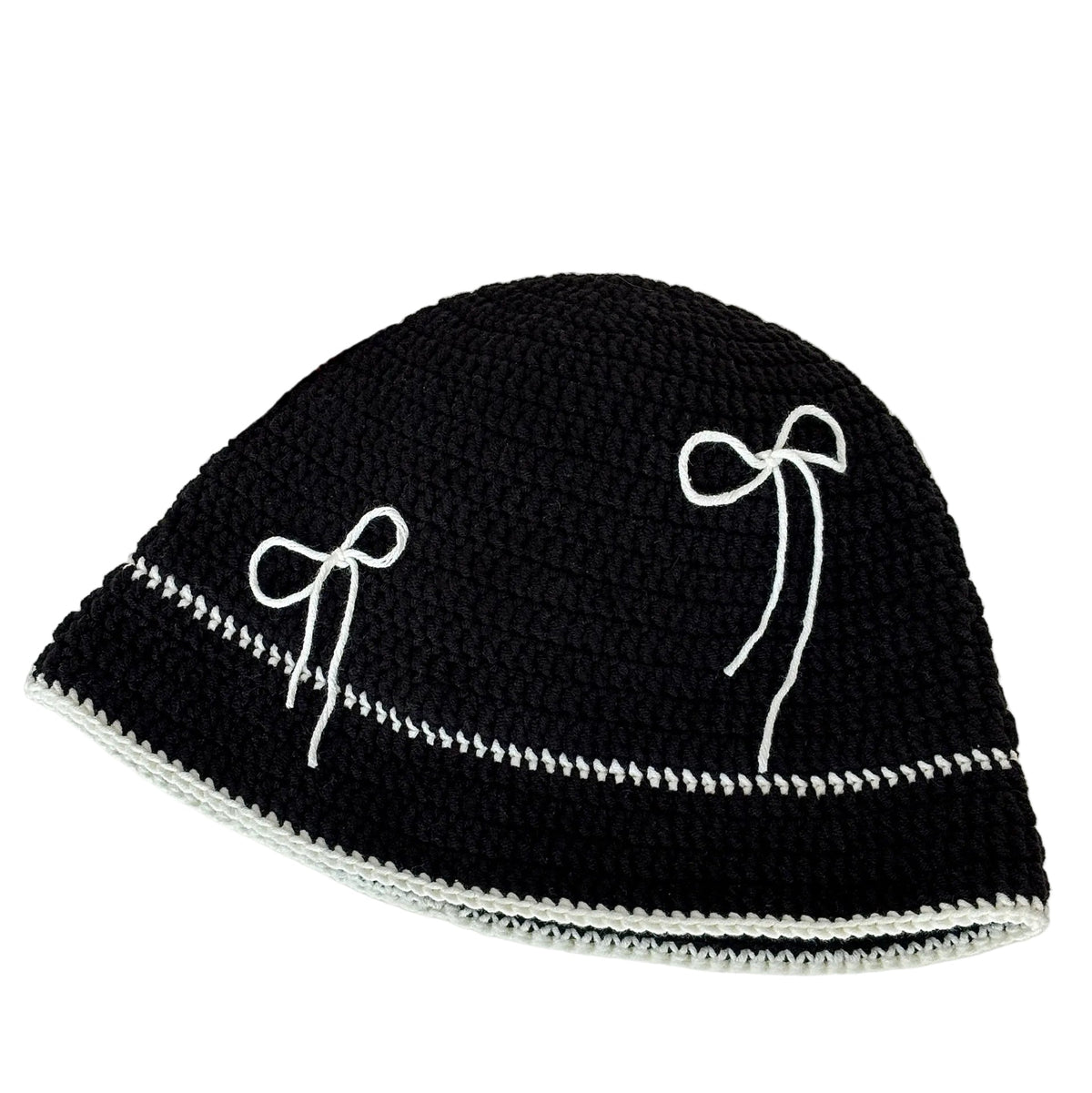 Black crochet hat with white bow patterns on a white surface, next to yarn and a newspaper.