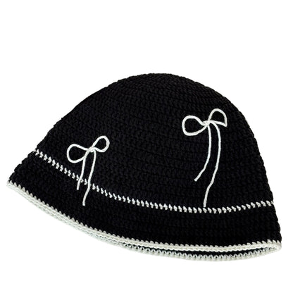 Black crochet hat with white bow patterns on a white surface, next to yarn and a newspaper.