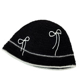 Black crochet hat with white bow patterns on a white surface, next to yarn and a newspaper.