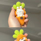 Hand holding a small, colorful carrot-shaped plush toy with a face on a gray background.