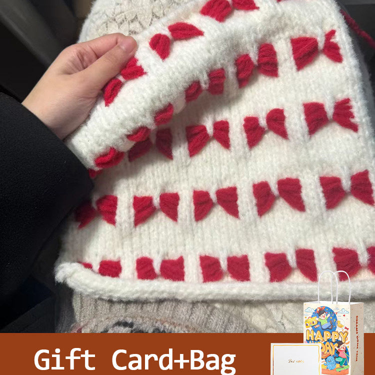 White knitted blanket with red patterns held by a hand, with a gift card and bag in the corner.